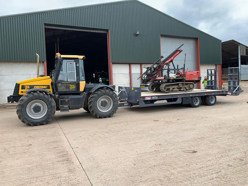 A large yellow and black JCB tractor pulling a flatbed trailer with a red drilling rig on it