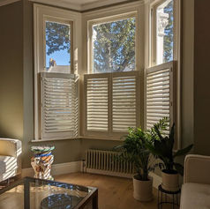 Bay window with plantation shutters a coffee table and house plants on a wooden floor