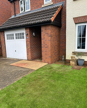 A garage with white doors and a brick exterior is adjacent to a small patch of grass and a paved driveway