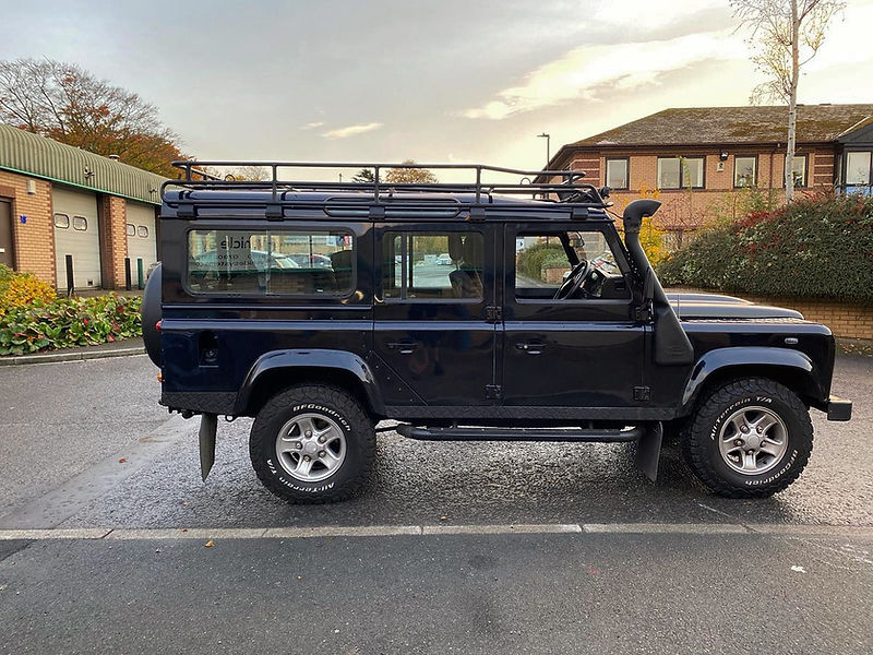 A dark blue Land Rover Defender 110 parked on tarmac, featuring a roof rack, snorkel, and off-road tyres
