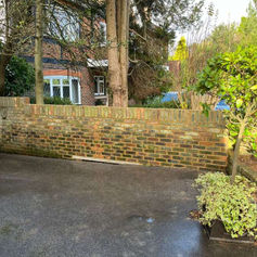 Long brick wall separating a tarmac driveway from a garden with trees and shrubs in front of a house