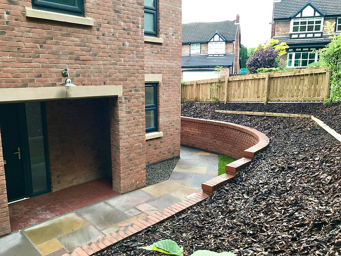 Curved brick retaining wall alongside a stone patio path leading to a home entrance with wood bark borders