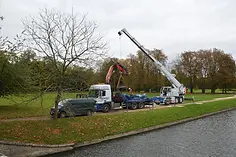 A crane on a truck lifts a heavy object near a riverbank, surrounded by autumn trees