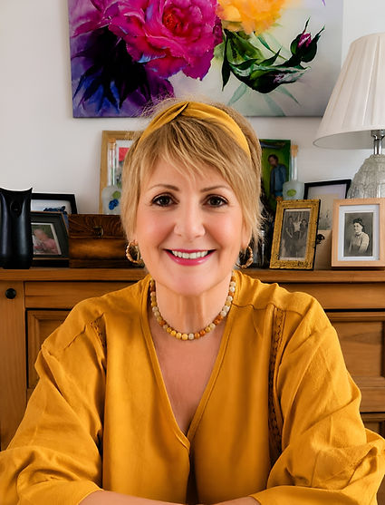 A smiling woman with blonde hair wearing a mustard yellow top sitting in front of bookshelves