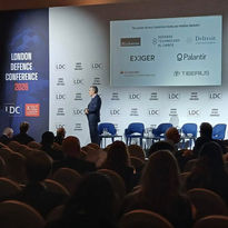 A speaker presenting on a stage during the London Defence Conference to an audience.