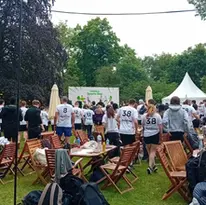 An outdoor gathering of people in white shirts with tables and umbrellas on grass.