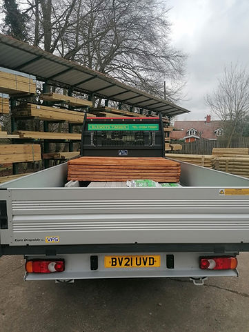 A flatbed truck loaded with wooden planks and bags, parked near timber shelves