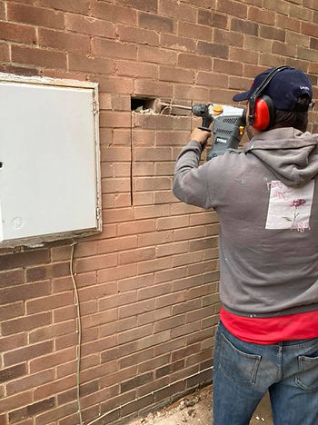 A person wearing a cap, earmuffs, and hoodie uses a power drill on a red brick wall