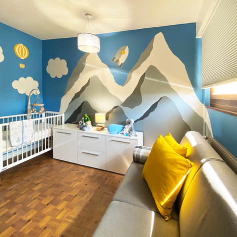 Baby room with wall mural illustrating mountains, clouds, rocket, airplane, hot balloon,  blue sky. Also showing a white crib, gray sofa and yellow pillows. Some toys and a lamp are placed on a dressing.   