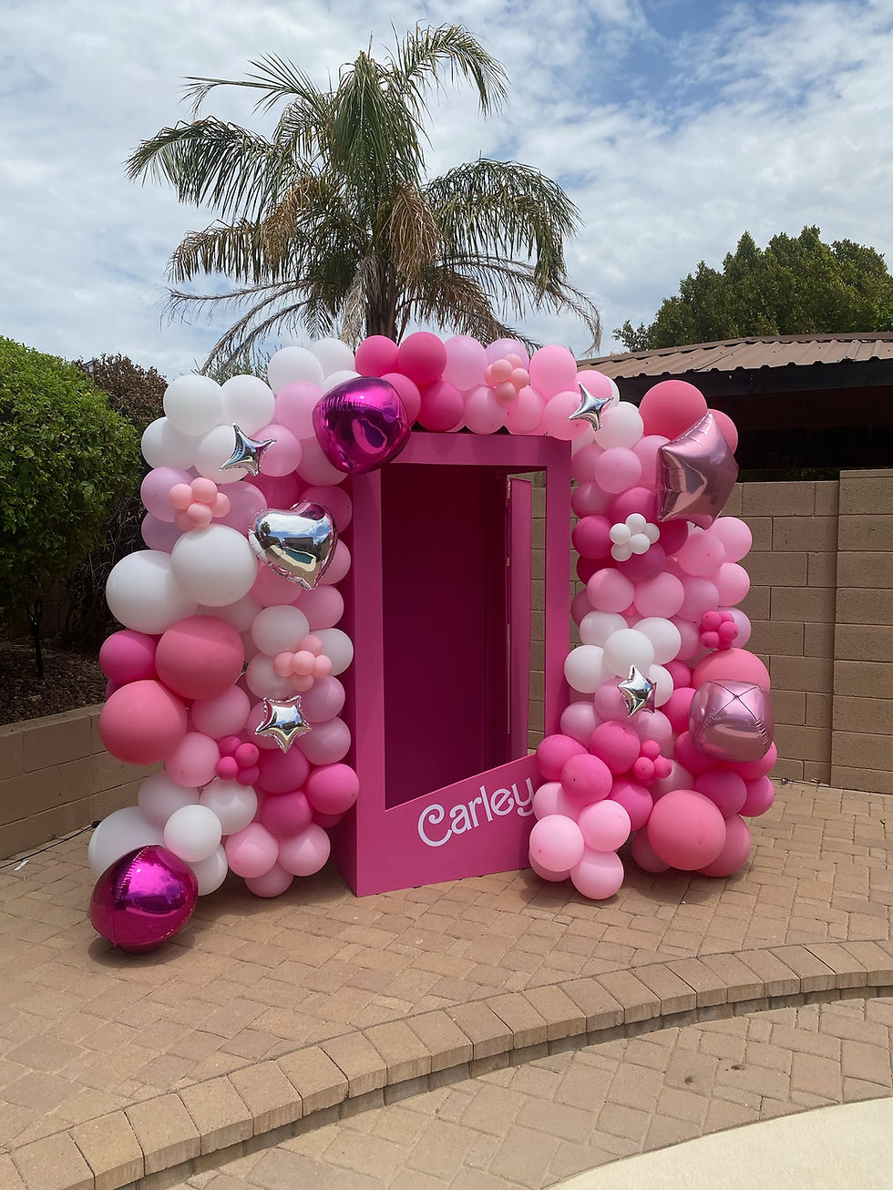 Pink and white balloon arch with metallic accents surrounds a pink door in an outdoor setting. The name "Carley" is written on the base.