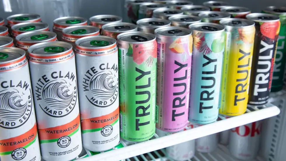 Cans of White Claw and Truly hard seltzers lined up on a refrigerator shelf. Varied colors and flavors visible, creating a vibrant display.