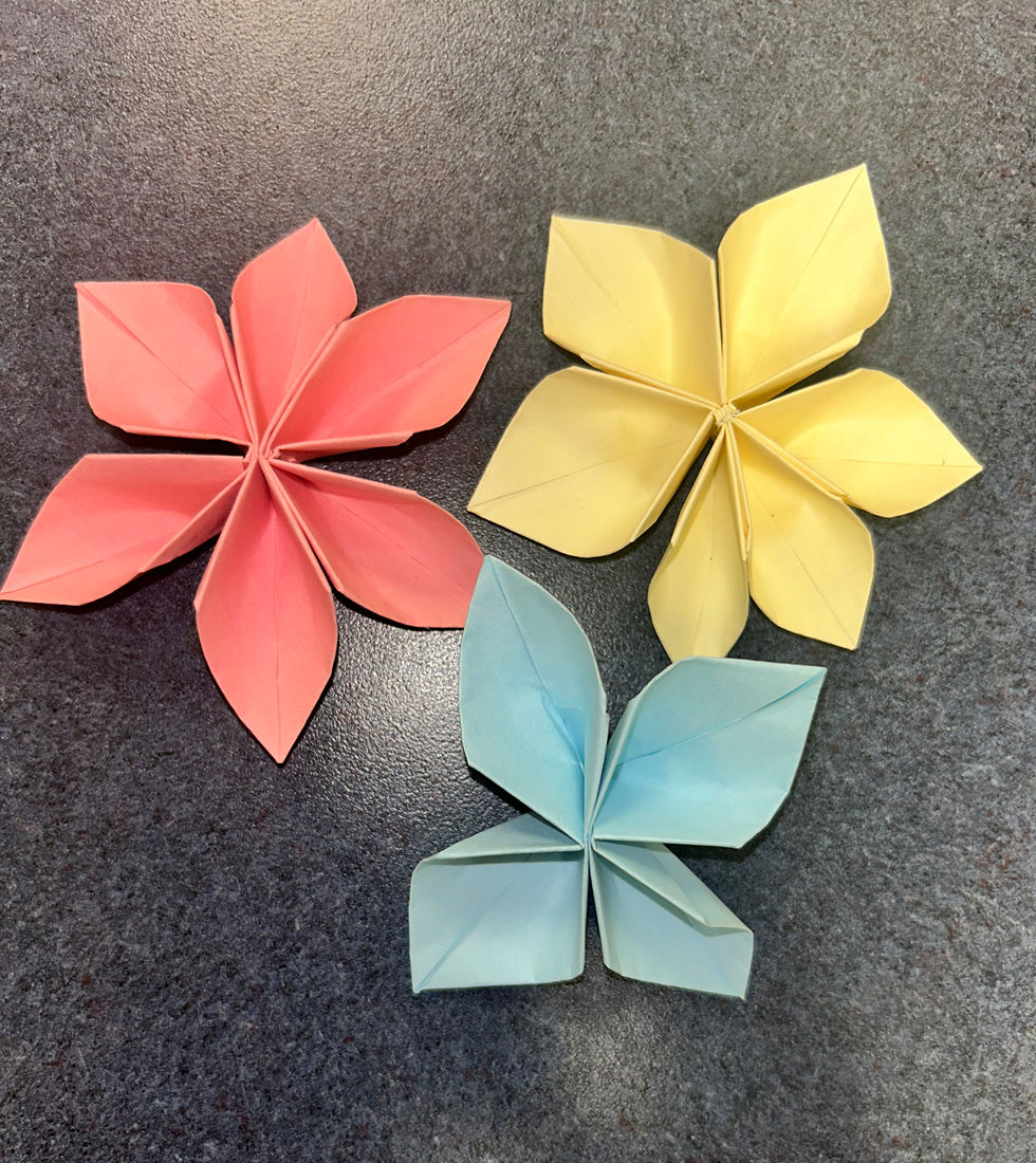 Three folded origami flowers in pink, yellow, and blue on a dark, textured surface. 