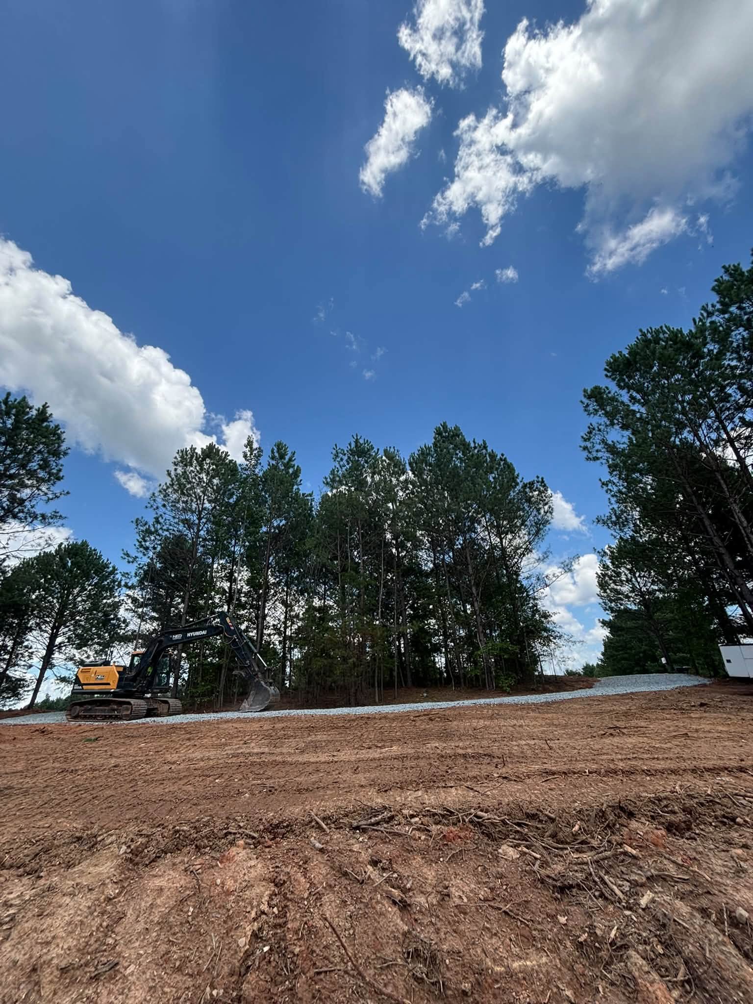 Site preparation with graded pad and new gravel driveway, excavator in background