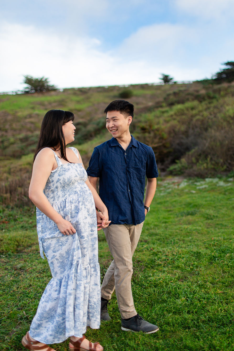 A couple holding hands and smiling, walking on green grass. The woman wears a blue patterned dress, the man in a navy shirt. Scenic hillside.
Sunset Spring Beach Maternity Session at Rockaway Beach in Pacifica by HKCreate Bay Area Photographers