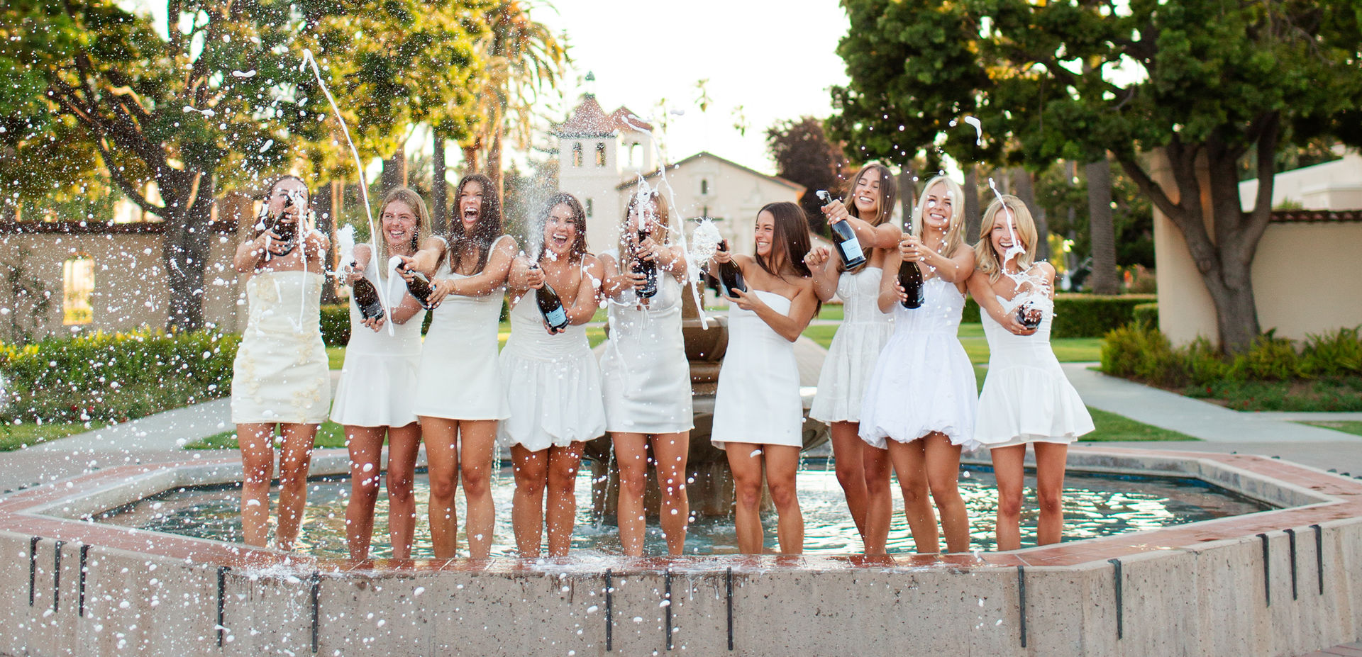 group of graduates celebrating in white dresses with champagne in a fountain
