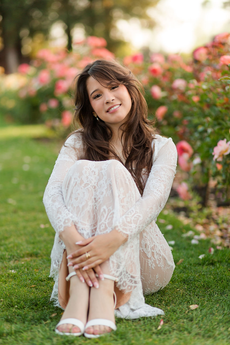 Young woman in a white lace dress sits on grass, smiling in a flower garden with pink blooms. San Jose Municipal Rosegarden Golden Hour College Graduation Photoshoot by Bay Area Photographers HKCreate
