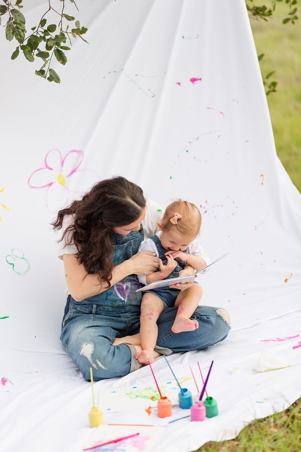 Mom and toddler painting on a white sheet outdoors. Both are in overalls, surrounded by colorful paints. Playful and creative mood.
Mommy and Me Maternity Outdoor Painting Session by HKCreate Bay Area Photographers at Guadalupe Oak Grove Park in San Jose