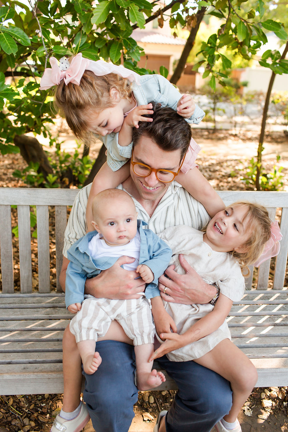 Smiling father sits on a bench with three young children outdoors under leafy trees, one child on his shoulders, warm family scene
Spring Family Photoshoot at Elizabeth Gamble Garden, Palo Alto by Bay Area Photographers HKCreate