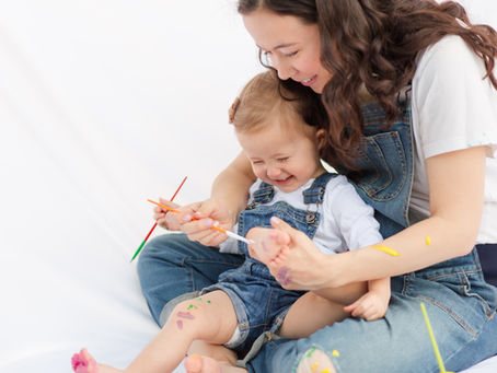 Mother and child joyfully paint each other in a playful setting. Both wear denim overalls with colorful paint on skin, against a white background. Happy and cute and playful Mommy and Me Maternity Painting Session by HKCreate Bay Area Photographers at Guadalupe Oak Grove Park in San Jose