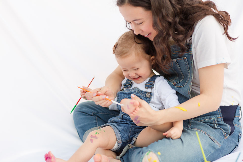 Mother and child joyfully paint each other in a playful setting. Both wear denim overalls with colorful paint on skin, against a white background. Happy and cute and playful Mommy and Me Maternity Painting Session by HKCreate Bay Area Photographers at Guadalupe Oak Grove Park in San Jose