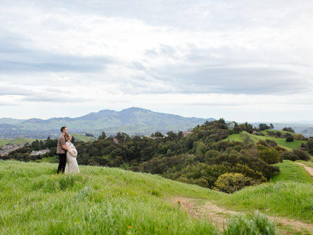 Couple embraces on a grassy hill with mountainous landscape in the background. Cloudy sky, lush greenery, serene and romantic vibe. Windy Green Hills Spring Maternity Session Briones Regional Park, Costa County East Bay by Bay Area Photographers HKCreate.