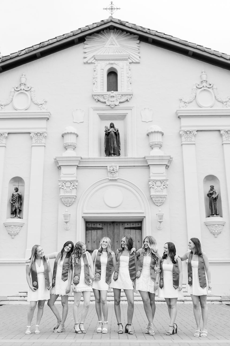 Seven women in white dresses and sashes pose smiling in front of the Mission at Santa Clara University in California. Fun Group Grad Photoshoot by Bay area Photographers HKCreate