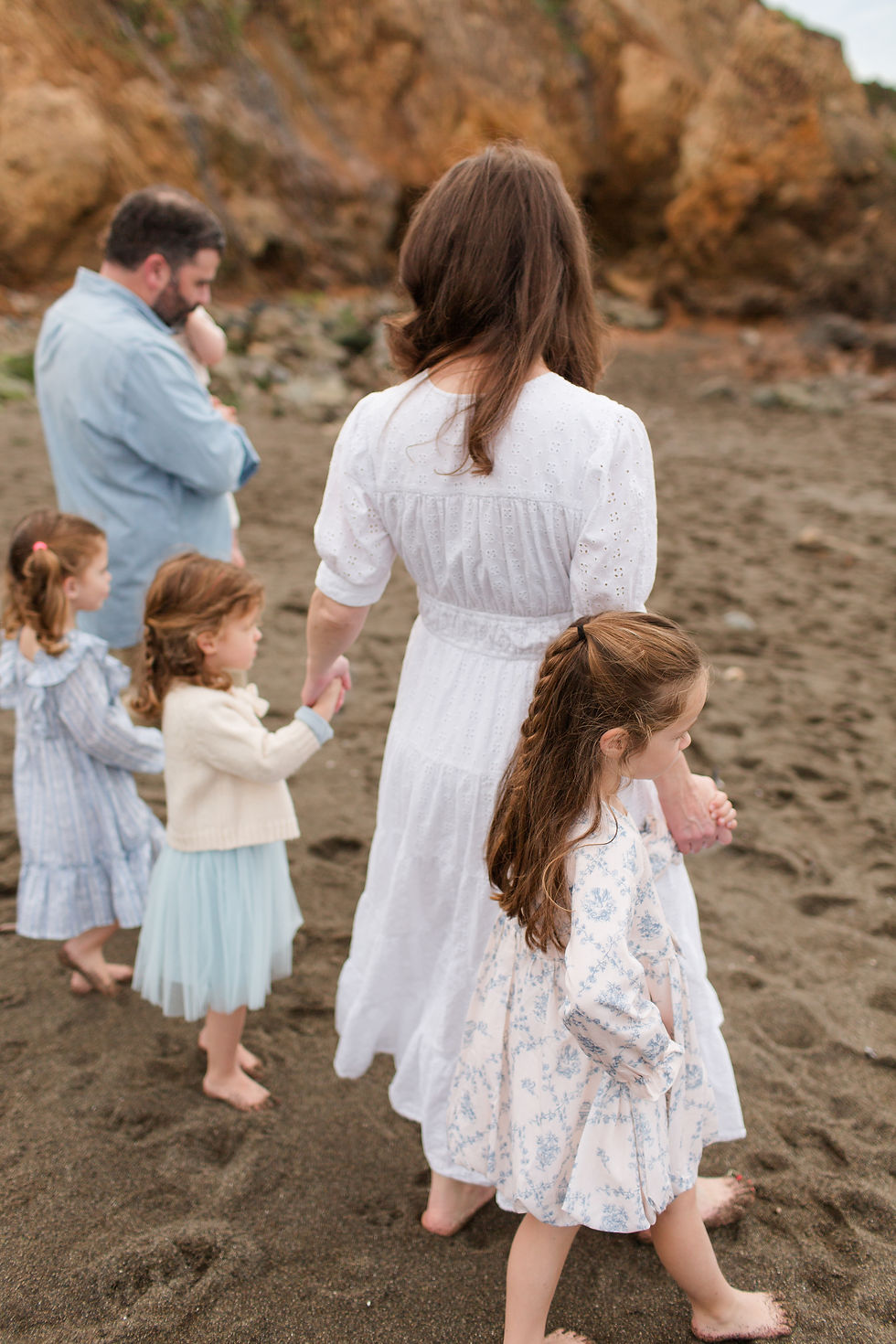 Family of six holding hands on a rocky beach, seen from behind, with a woman in white and children in pastel dresses. Overcast Beach Family Photoshoot at Rockaway Beach in Pacifica by Bay Area Photographers HKCreate
