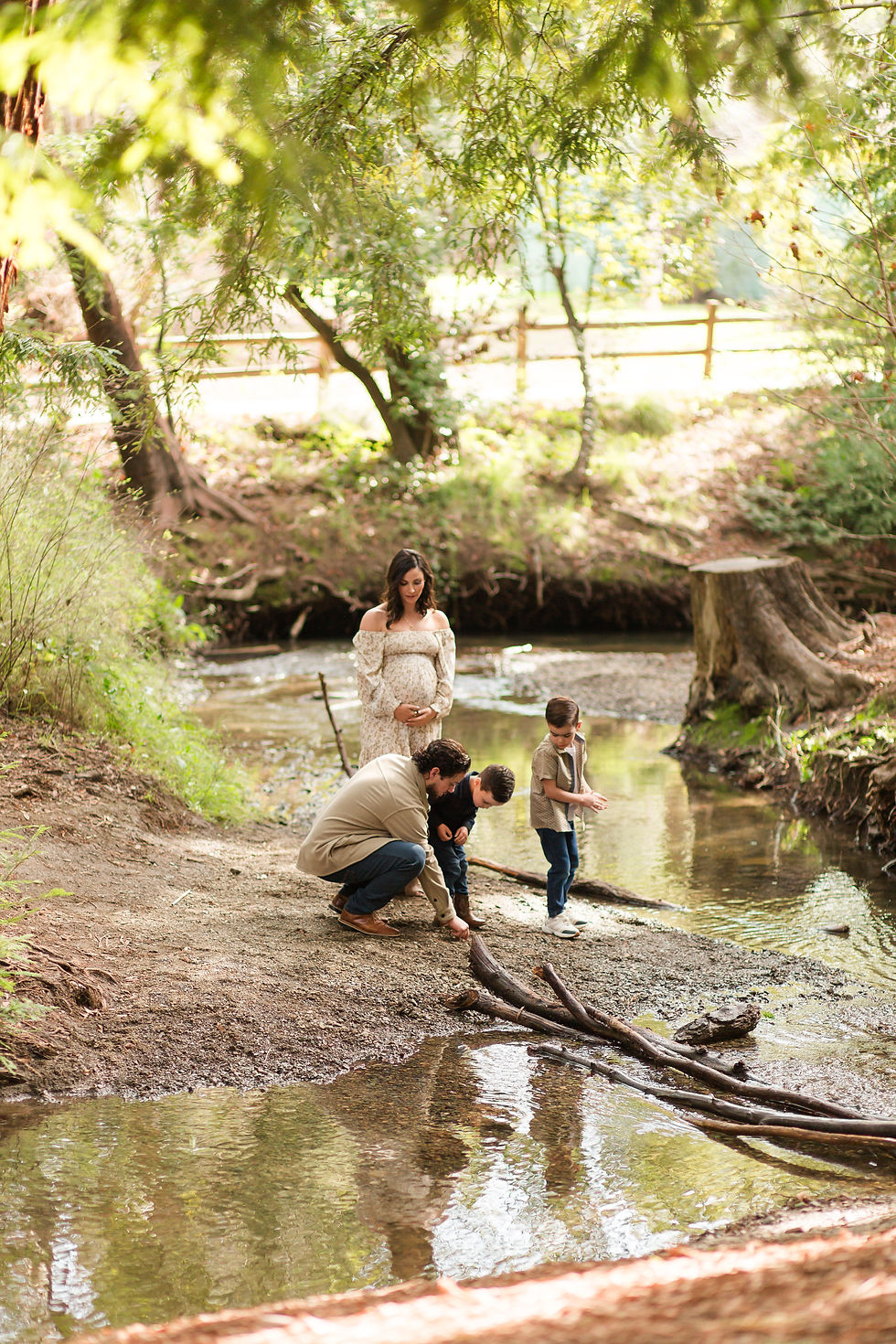 Pregnant woman stands by a creek as a dad and this two sons crouch near the water in a sunny wooded area.