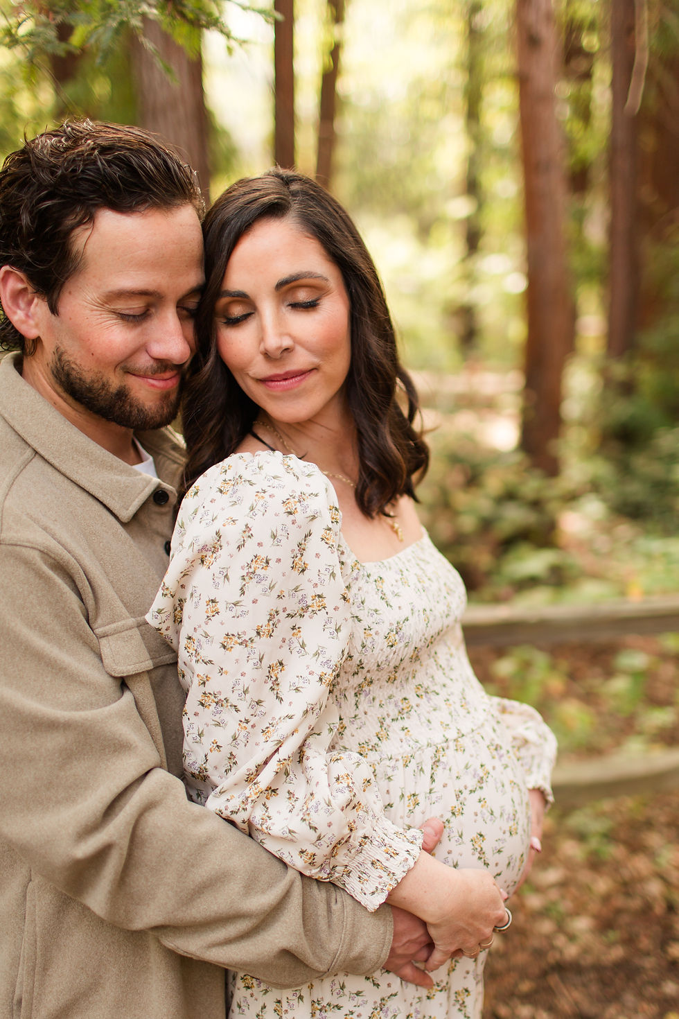 Pregnant woman in a floral dress embraced by a man in a beige jacket in a sunlit forest, both eyes closed in a tender pose. Winter Family Maternity Photoshoot at Redwood Grove Nature Preserve, Los Altos by Bay Area Photographers HKCreate