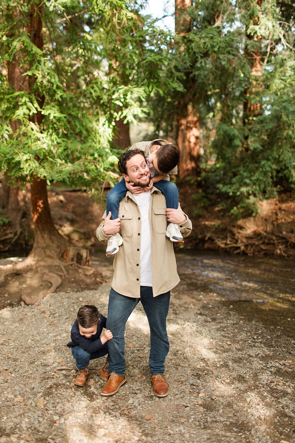 Smiling man carries a child on his shoulders while another hugs his leg in a sunlit forest by a creek. Winter Family Maternity Photoshoot at Redwood Grove Nature Preserve, Los Altos by Bay Area Photographers HKCreate