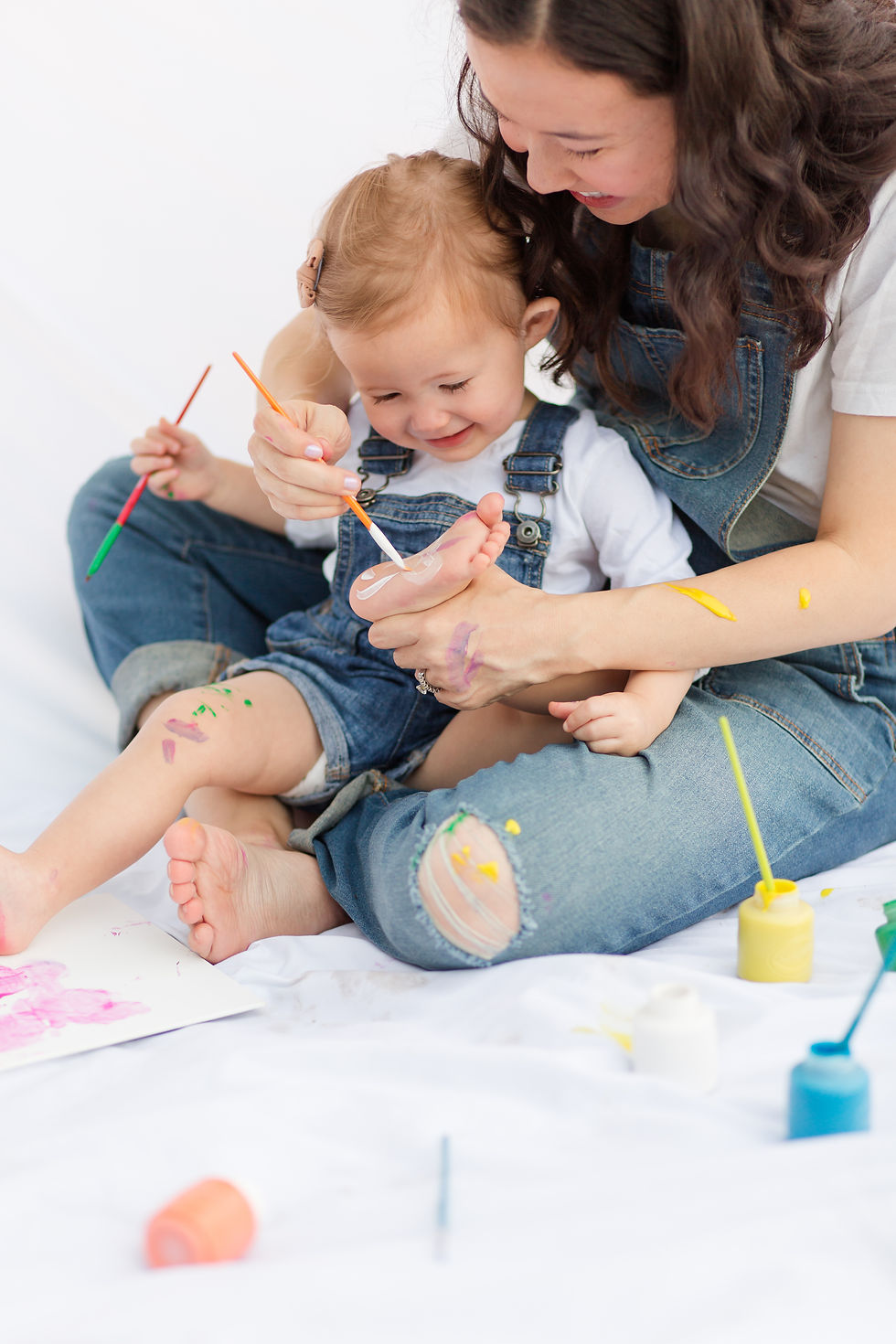 Mother and child in denim overalls, painting on feet with colorful brushes. Bright paints and a playful, creative mood on a white background.
Mother and child joyfully paint each other in a playful setting. Both wear denim overalls with colorful paint on skin, against a white background.
Happy and cute and playful Mommy and Me Maternity Painting Session by HKCreate Bay Area Photographers at Guadalupe Oak Grove Park in San Jose