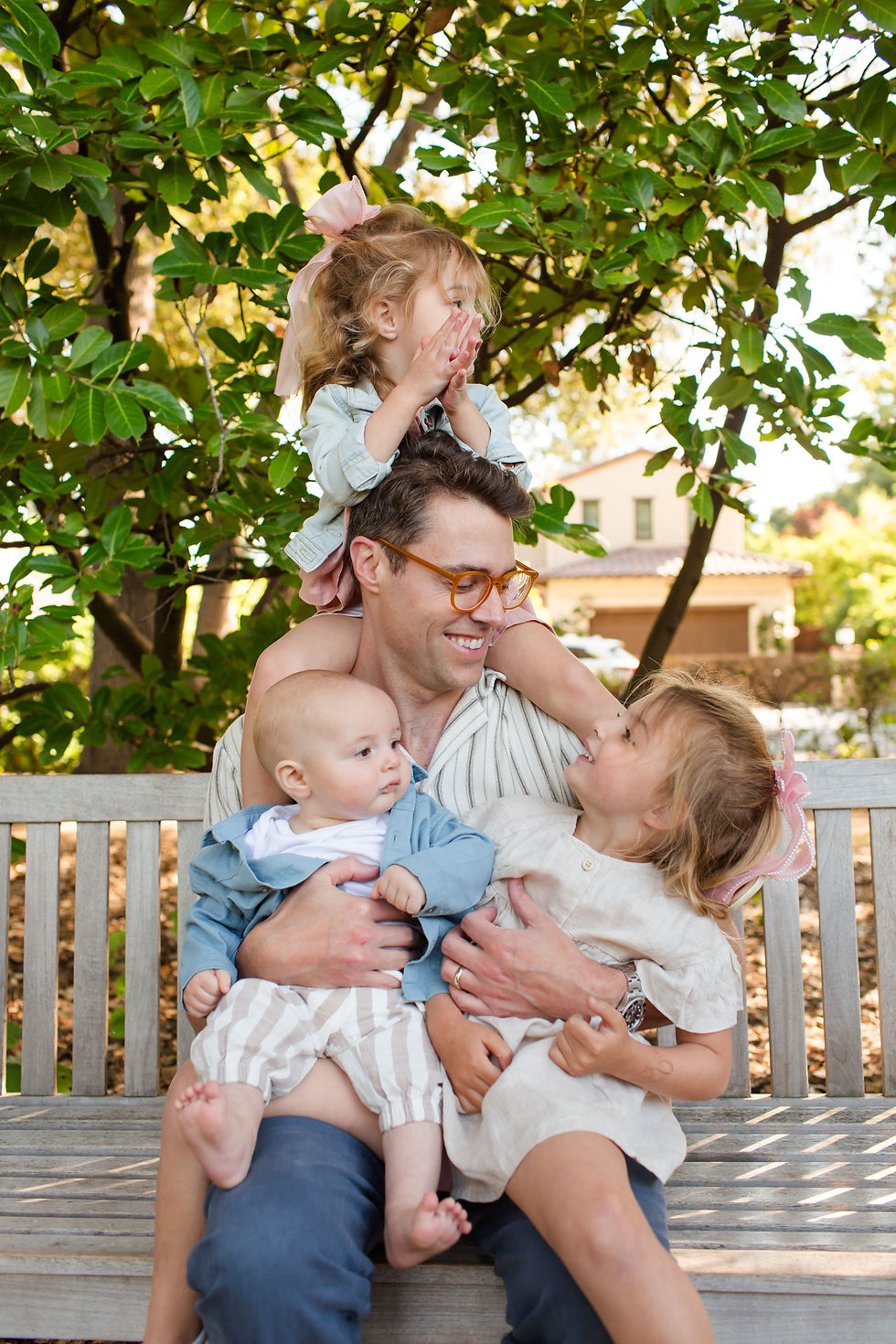 Smiling father sits on a bench with three young children in a leafy yard, one girl on his shoulders, another cuddling him, baby in arms
Spring Family Photoshoot at Elizabeth Gamble Garden, Palo Alto by Bay Area Photographers HKCreate