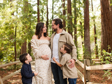 Pregnant couple and two young boys smiling and hugging on a wooden forest path among tall redwood trees. Winter Family Maternity Photoshoot at Redwood Grove Nature Preserve, Los Altos by Bay Area Photographers HKCreate