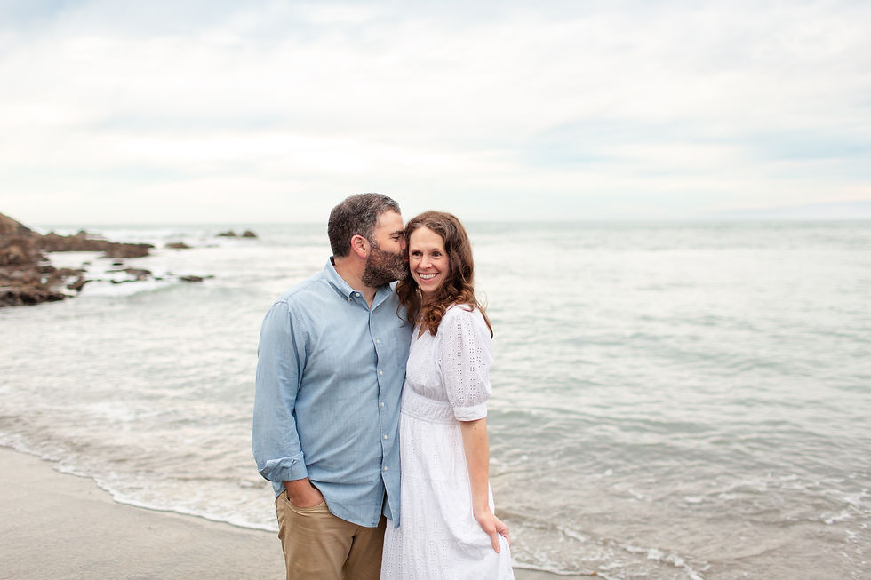 Couple on a rocky beach, man kissing woman’s temple as she smiles in a white dress, with calm ocean and cloudy sky behind. Overcast Beach Family Photoshoot at Rockaway Beach in Pacifica by Bay Area Photographers HKCreate