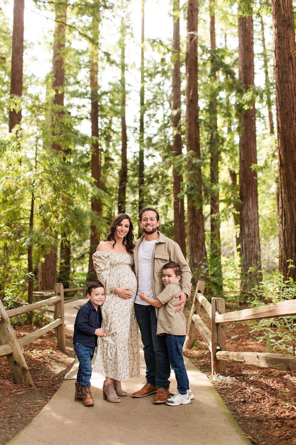 Pregnant woman and man with two young boys pose smiling on a forest path in tall redwoods. Winter Family Maternity Photoshoot at Redwood Grove Nature Preserve, Los Altos by Bay Area Photographers HKCreate