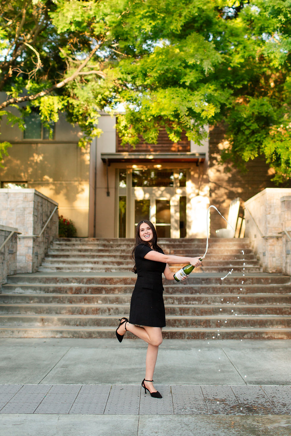 Smiling woman in a black dress pops a champagne bottle on stone steps under green trees, with spray flying.
College Grad photoshoot at Sonoma State University  by Bay Area Photographers HKCreate