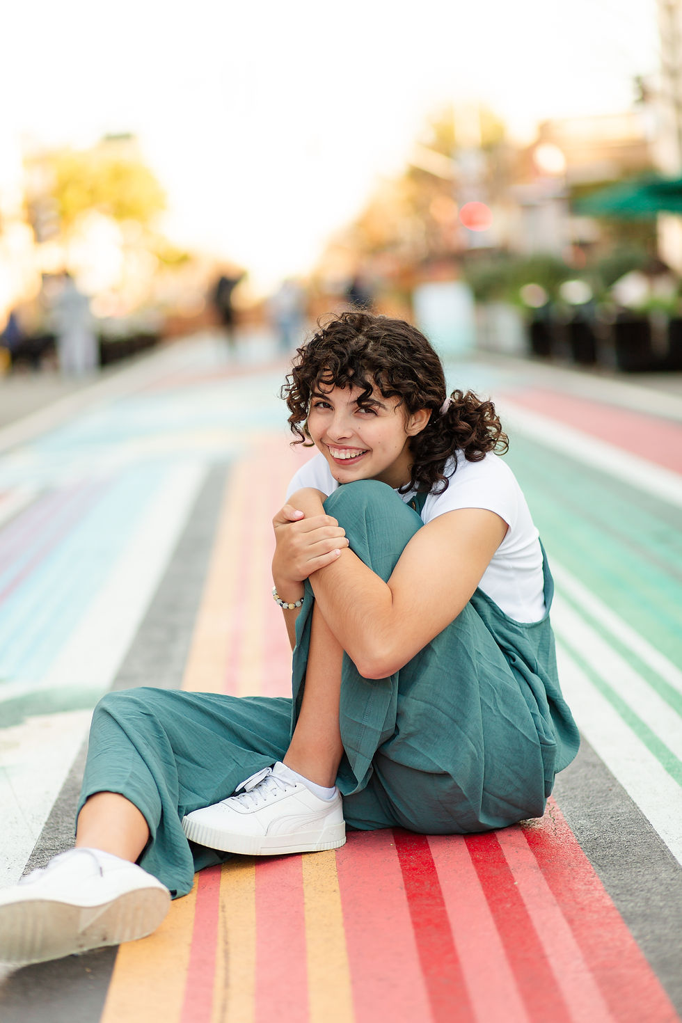 Teenage girl with curly hair sits on a vibrant, multicolored street. Wearing a white shirt and teal overalls, creating a joyful vibe.
Downtown San Mateo Senior High School Graduation Urban Photoshoot by Bay Area Photographers HKCreate
