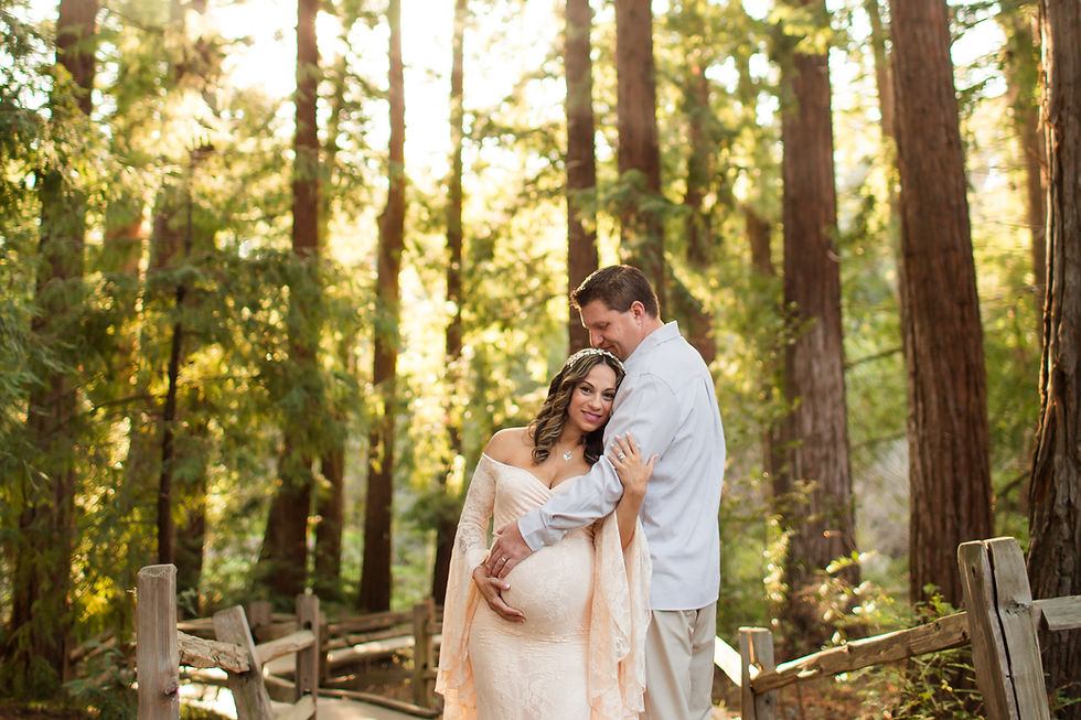 Man with his hand on his pregnant wife's belly. Woman wearing cream lace dress in a forest and man wearing neutral color outfit. Romantic glow behind them. Redwood Grove Nature Preserve Maternity Photoshoot in Los Altos by Bay Area San Jose Photographers HKCreate.