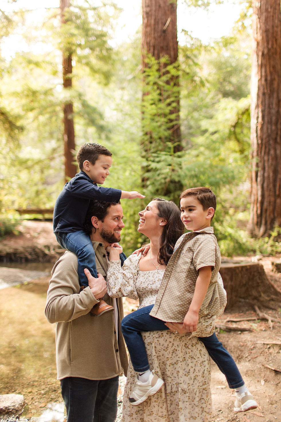 Smiling parents hold two young sons in a sunlit forest, one boy on dad’s shoulders reaching toward mom. Winter Family Maternity Photoshoot at Redwood Grove Nature Preserve, Los Altos by Bay Area Photographers HKCreate