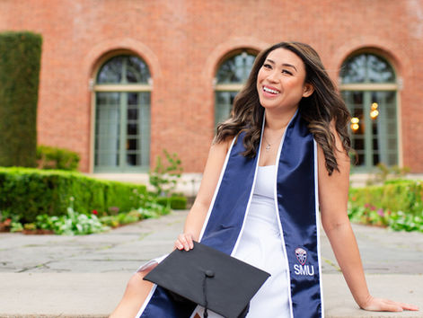 Smiling woman in white dress and blue SMU sash holds a graduation cap in a garden courtyard before a brick building. Samuel Merritt University Spring Grad Photoshoot by HKCreate Bay Area Photographers  Filoli Historic House and Garden, Woodside 
