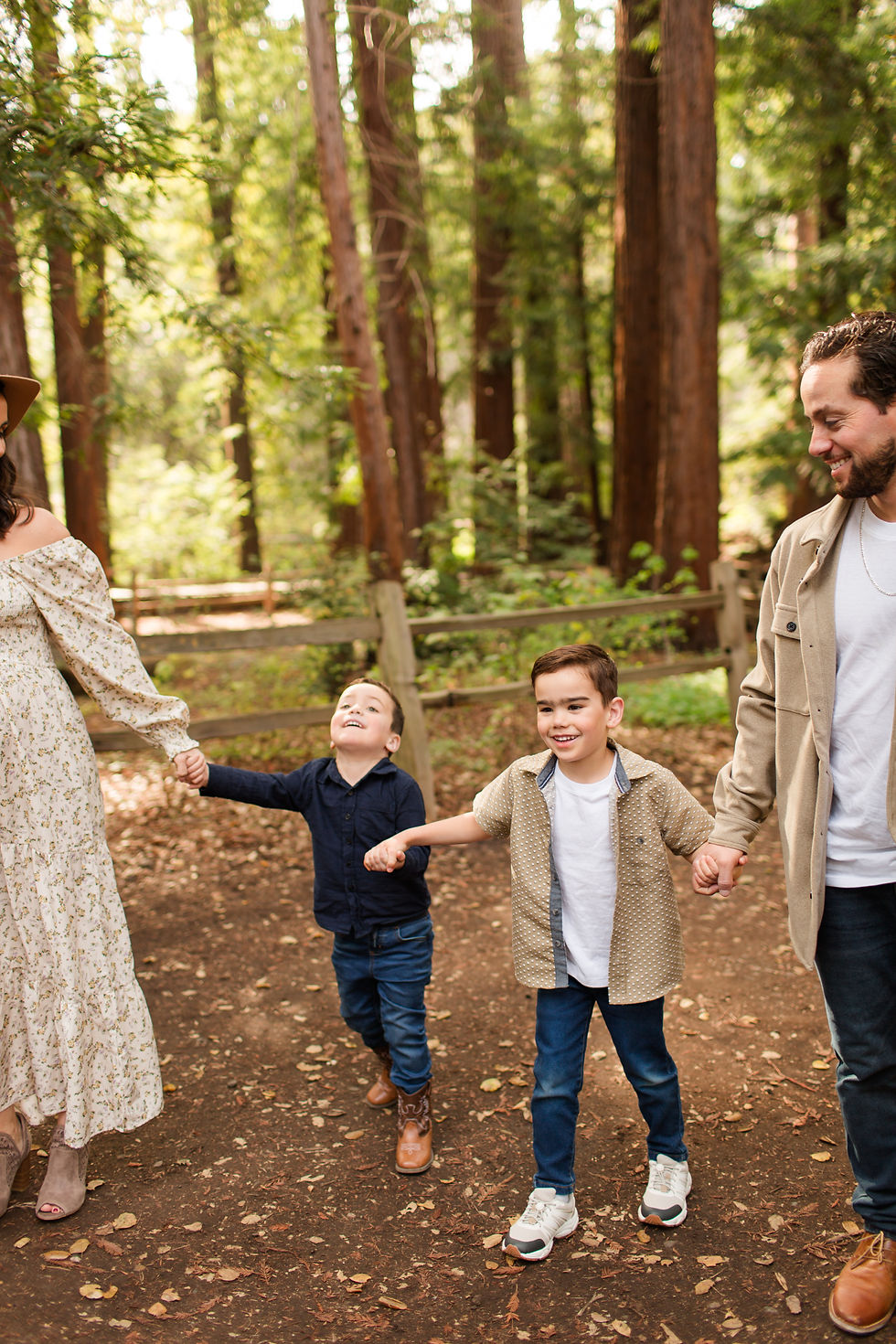 Family of four holding hands on a sunlit forest path, smiling and walking among tall trees and a wooden fence. Winter Family Maternity Photoshoot at Redwood Grove Nature Preserve, Los Altos by Bay Area Photographers HKCreate
