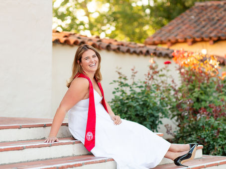 Smiling woman in a white dress with a red sash sits on tiled steps in a sunny courtyard with flowers and terracotta roofs. Mission steps at SCU Golden Hour Santa Clara University Graduation Photoshoot by Bay Area Photographers HKCreate