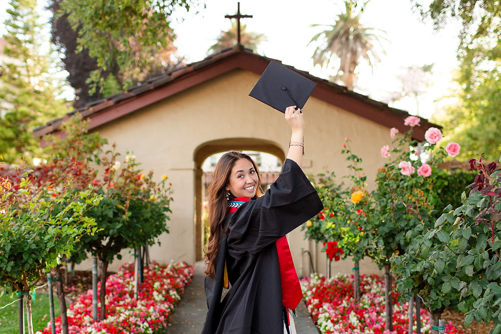 Smiling graduate in black gown raises a cap in a flower-filled garden at the Mission Gardens on the SCU Campus. Santa Clara University Grad Photoshoot by Bay Area Photographers HKCreate.