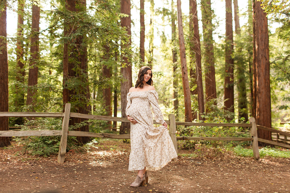 Pregnant woman in a white floral dress posing by a wooden fence in a sunlit redwood forest, holding her belly and smiling. Winter Family Maternity Photoshoot at Redwood Grove Nature Preserve, Los Altos by Bay Area Photographers HKCreate