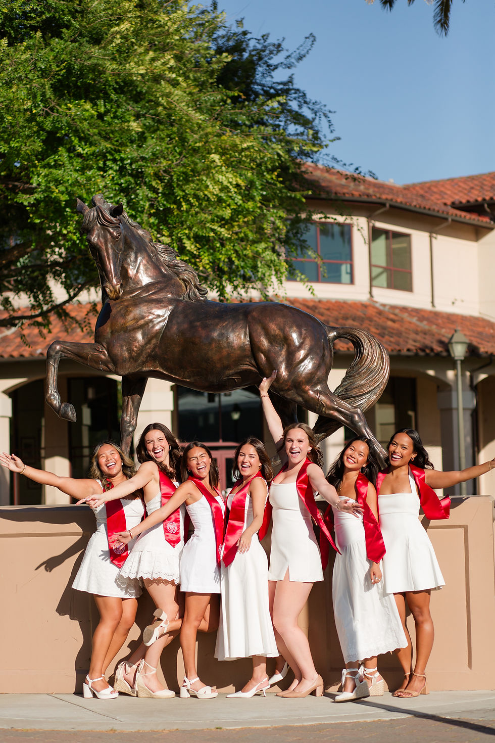 Seven smiling women in white dresses and red stoles pose by a bronco statue on a sunny campus. Fun Group Grad Photoshoot at Santa Clara University by Bay Area Photographers HKCreate