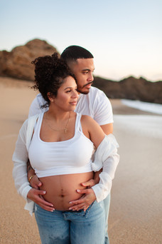 Sunset golden hour maternity beach photoshoot at Cowell Ranch State Beach in Half Moon Bay. Couple embracing each other hugging pregnant woman's belly. Serene and golden glow. Couple wearing neutral colored outfits. Pregnant woman wearing white top with jeans and open shirt showing belly
