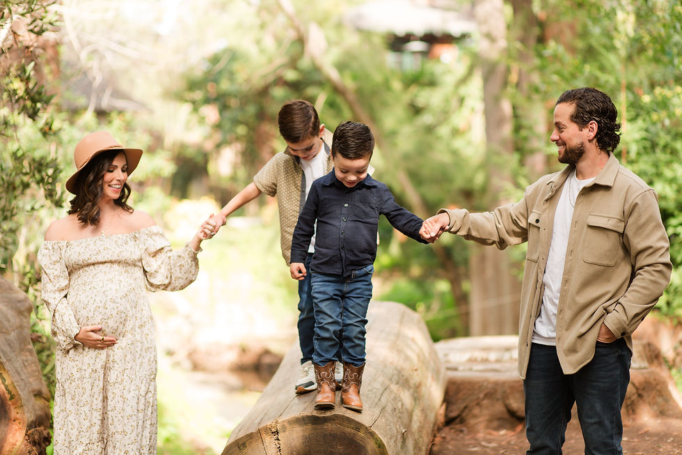 Pregnant woman and man hold hands with two boys balancing on a log in a sunlit forest, all smiling warmly. Winter Family Maternity Photoshoot at Redwood Grove Nature Preserve, Los Altos by Bay Area Photographers HKCreate