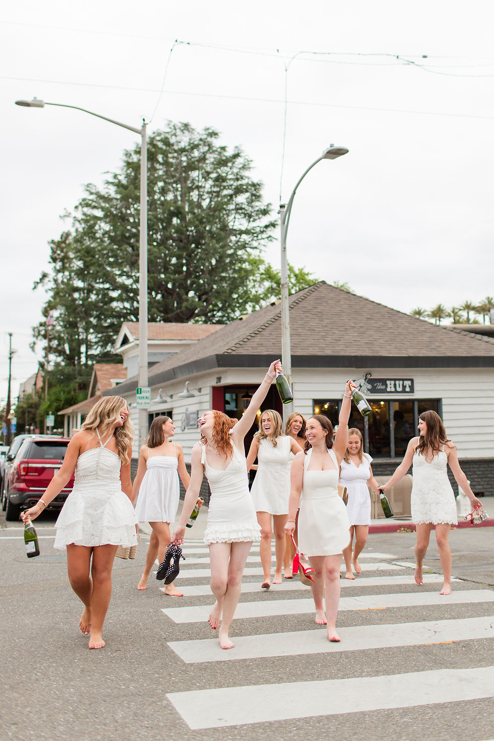 Eight women in white dresses walk barefoot across a crosswalk, holding shoes and champagne bottles in front of The Hut campus restaurant. Santa Clara University fun group grad photoshoot by bay area photographers HKCreate