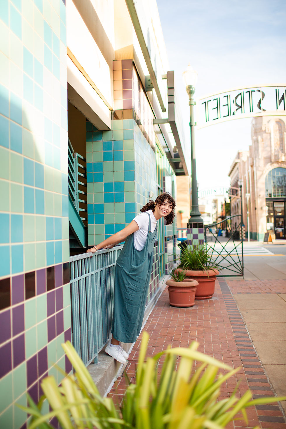 Smiling person leans over a railing beside a colorful tiled building. Potted plants line the brick walkway. Bright, sunny day.
Downtown San Mateo Senior High School Graduation Urban Photoshoot by Bay Area Photographers HKCreate