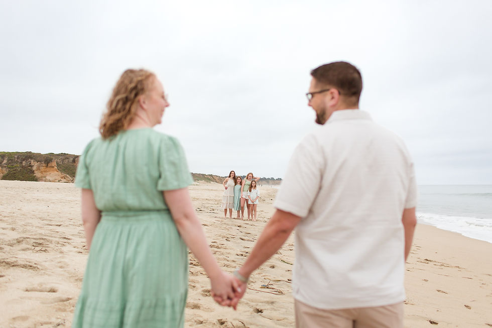 Couple holding hands on a beach, looking at a smiling family of four in the distance under a cloudy sky. Summer family photoshoot at Half Moon Bay State Beach in California by Bay Area Photographers HKCreate
